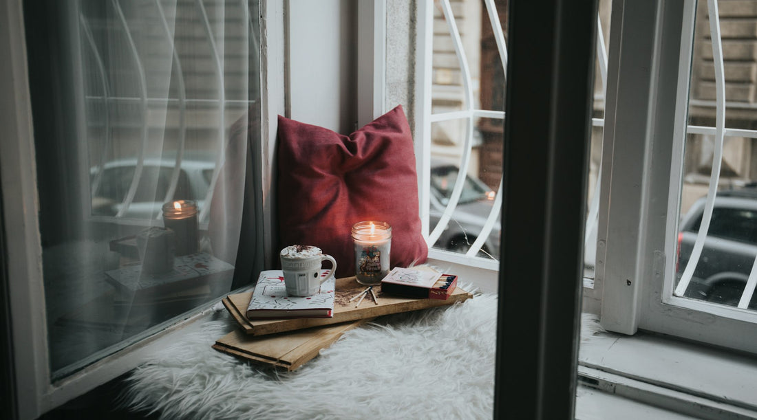 Cosy window nook with a lit candle, a warm mug and cushions, the setting for a winter evening wind-down routine