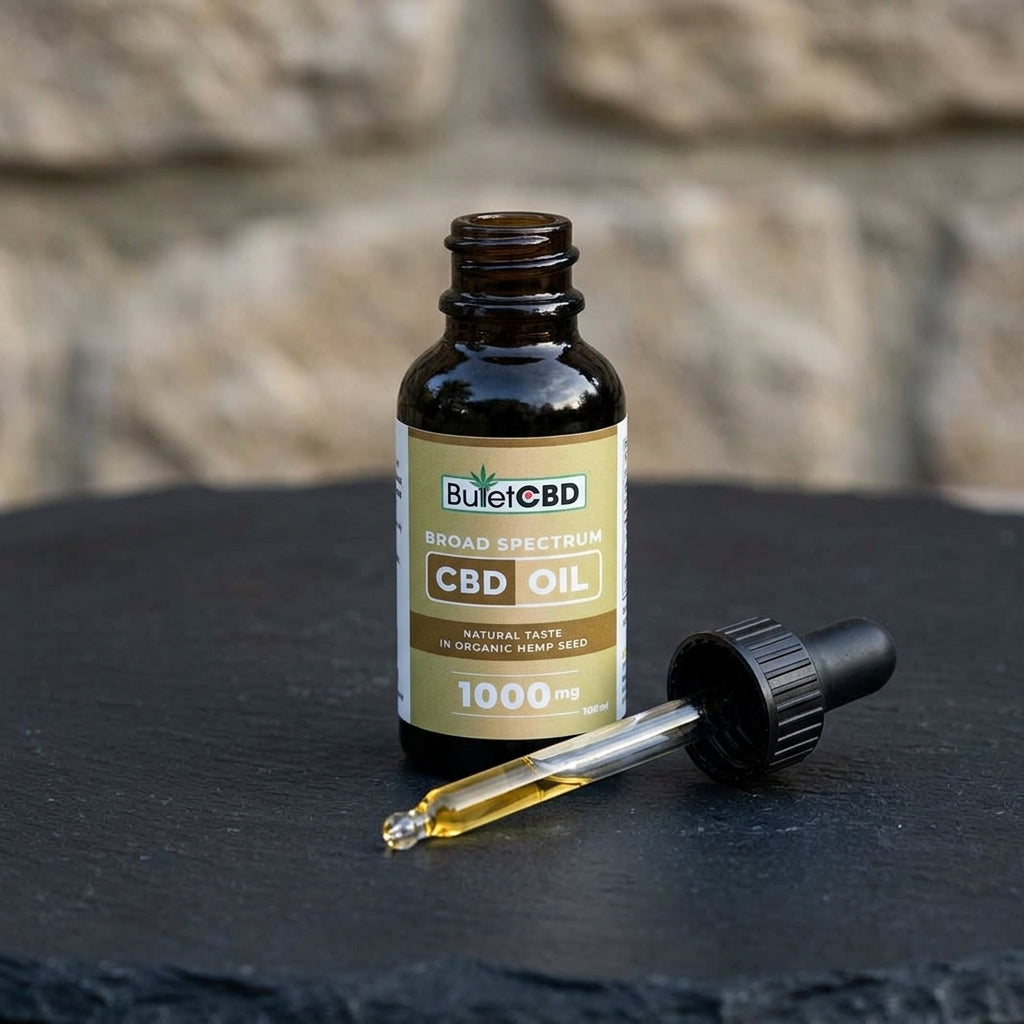 A bottle of BulletCBD Broad Spectrum CBD Oil next to a glass dropper containing amber oil, set on a dark slate surface with a blurred stone wall background.