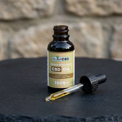 A bottle of BulletCBD Broad Spectrum CBD Oil next to a glass dropper containing amber oil, set on a dark slate surface with a blurred stone wall background.
