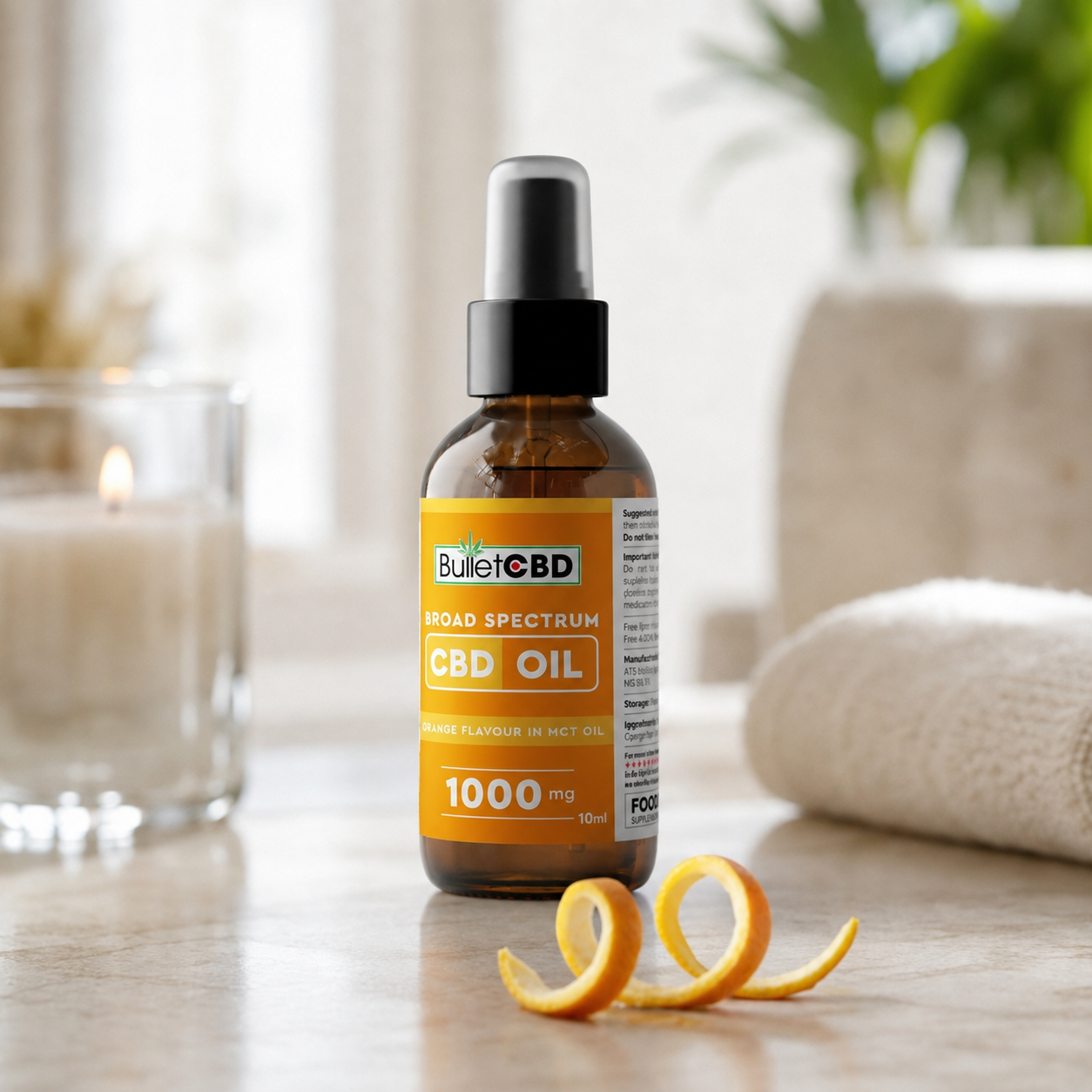 A 10ml amber glass bottle of BulletCBD Broad Spectrum CBD Oil with a black dropper cap, styled on a wooden surface with a curled orange peel and a blurred background featuring a candle, towel, and plant.