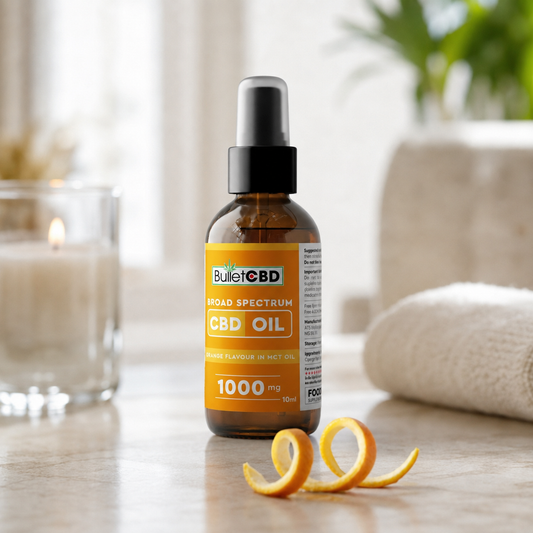 A 10ml amber glass bottle of BulletCBD Broad Spectrum CBD Oil with a black dropper cap, styled on a wooden surface with a curled orange peel and a blurred background featuring a candle, towel, and plant.